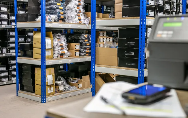 A dispatch area with picking shelves, cartons and poly-bagged stock beside a label printer and handheld scanner on the packing bench