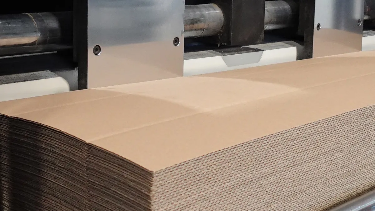 A stack of plain unprinted kraft corrugated blanks in front of the print and slotting units of a converting line