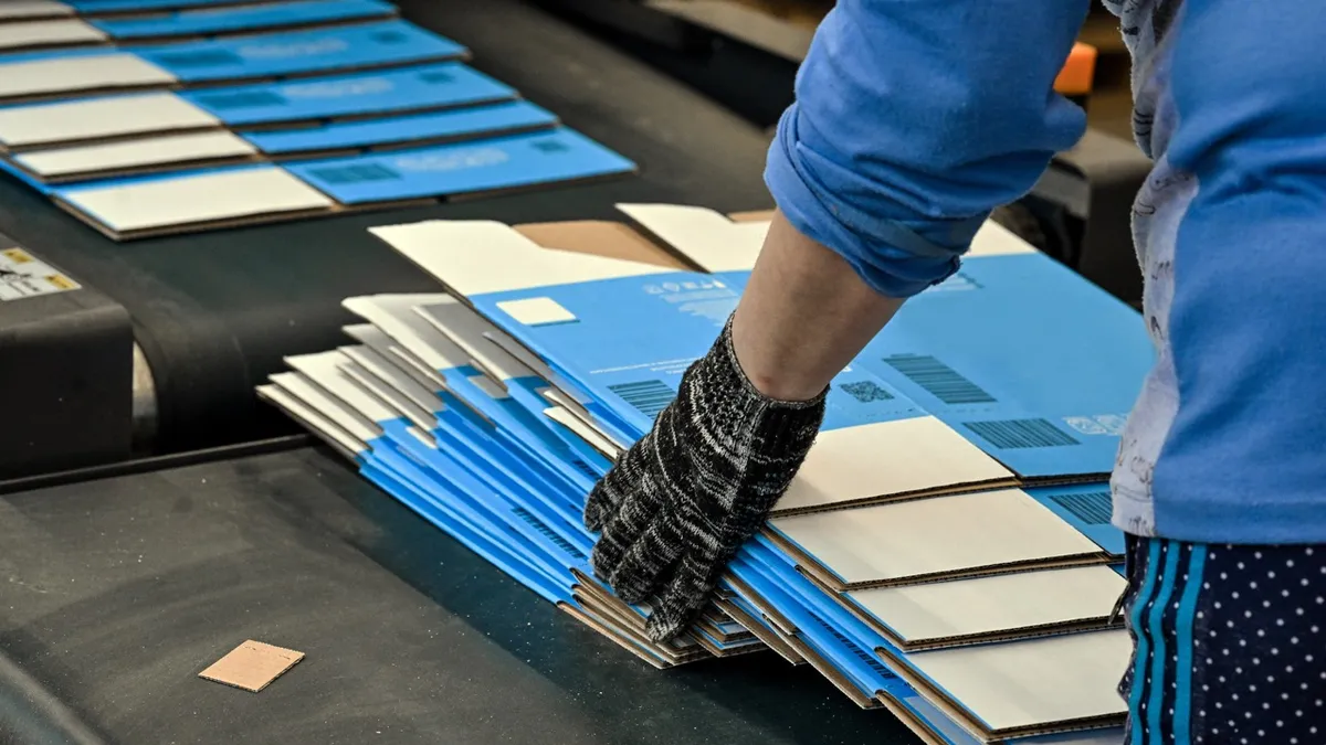 A gloved hand steadying a stack of blue and white printed corrugated blanks on a converting line