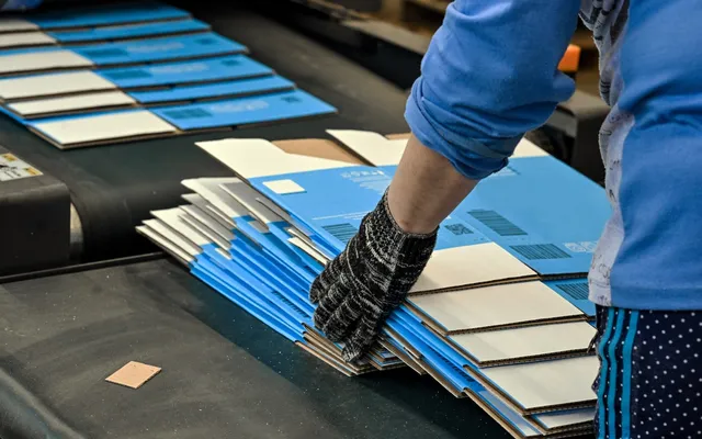 A gloved hand steadying a stack of blue and white printed corrugated blanks on a converting line